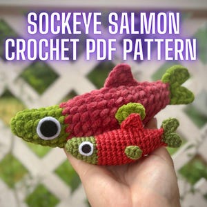 May include: Two handmade crochet sockeye salmon toys in different sizes, featuring deep red bodies, green heads, and green tails. Each fish has a white and black circular eye. The text SOCKEYE SALMON CROCHET PDF PATTERN is displayed above.