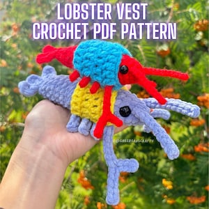 May include: Two crocheted lobster toys, one blue and yellow, the other purple, with red claws and black eyes. The toys are held in a hand against a background of green foliage.