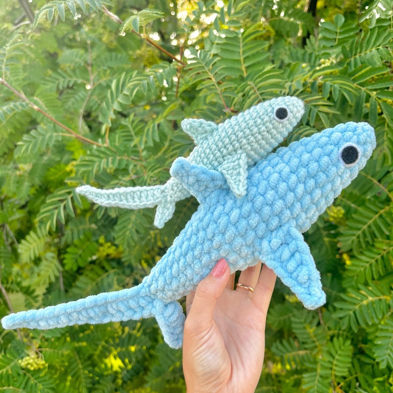 Pu&ograve; includere: Due peluche di squalo blu all'uncinetto. Lo squalo pi&ugrave; piccolo &egrave; appollaiato sopra quello pi&ugrave; grande. Entrambi gli squali hanno gli occhi neri.