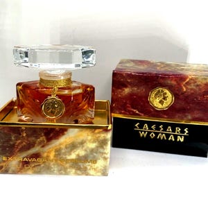 May include: A gold and red box with the text "CAESARS WOMAN" on the front. The box contains a clear glass bottle of perfume with a gold cap and a gold coin charm. The bottle is labeled "EXTRAVAGANT PERFUME 1/2 FL OZ 15 ml."