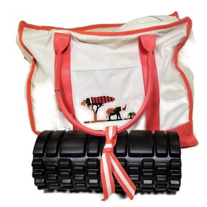 May include: A white canvas tote bag with a coral trim and a small embroidered design of a tree and elephants. The bag is sitting on top of a black foam roller with a coral and white ribbon.
