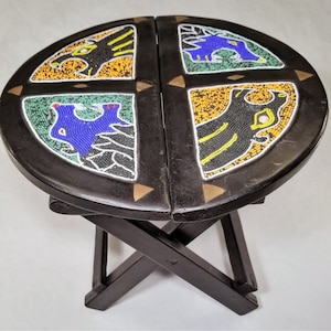 May include: A black wooden folding stool with a round seat decorated with colourful beaded designs. The designs feature geometric patterns and stylised animal figures.