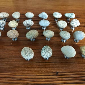May include: A collection of 20 natural stone drawer pulls in various shades of grey, brown, and white. Each pull is a unique, irregularly shaped stone with a silver metal base.