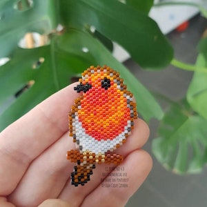 Puede incluir: Un broche de abalorios con forma de petirrojo, con pecho rojo, naranja y blanco, alas marrones y ojos negros. El pájaro está posado en una rama marrón. El texto "CC BY-NC-ND 4.0" es visible.