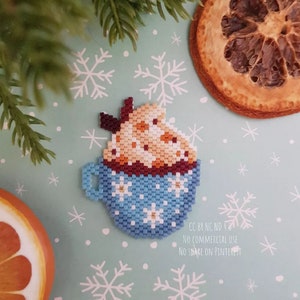 Peut inclure: Une tasse bleue et blanche avec un bord brun et des flocons de neige blancs. La tasse est remplie d'un liquide brun surmonté de crème fouettée blanche et de pépites de chocolat. L'arrière-plan est bleu clair avec des flocons de neige blancs.