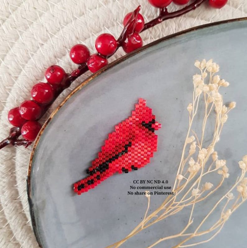 Puede incluir: Un broche de p&aacute;jaro cardenal rojo con detalles negros, hecho con cuentas, sobre un plato gris. El ave est&aacute; elaborada con peque&ntilde;as cuentas, mostrando un dise&ntilde;o detallado. Tambi&eacute;n se ven bayas rojas y flores secas.