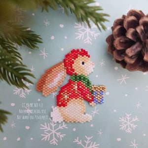 Puede incluir: Un adorno navideño de cuentas de un conejito que lleva un atuendo rojo y verde y sostiene una caja de regalo azul y blanca. El conejito está sobre un fondo azul claro con copos de nieve blancos.