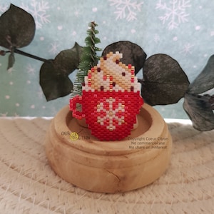 Peut inclure: Une tasse rouge en perles avec un flocon de neige blanc et une garniture de crème fouettée blanche en perles. La tasse est posée sur une base en bois avec un petit sapin de Noël vert derrière elle. Le texte "Copyright Coeur Citron No commercial use No share on Pinterest" est visible sous la tasse.