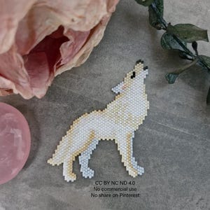 May include: A beaded white wolf figurine in a howling pose. The wolf is made of small, square beads in shades of white and cream. The figurine is set against a gray background with a pink flower and a pink crystal ball.