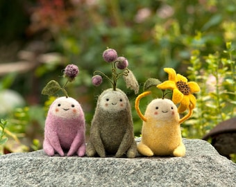May include: Three felt figures with smiling faces and plant-like features. The figures are pink, green, and yellow and are sitting on a gray rock. The pink figure has a purple flower on its head, the green figure has a green flower on its head, and the yellow figure has a yellow flower on its head.