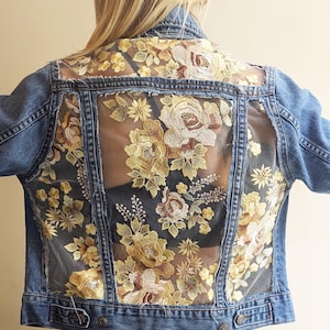 以下が含まれることがあります： イエローとクリーム色の花の刺繍が施された、シースルーのバックパネルが付いたデニムジャケット。 ジャケットは、クラシックな襟、ボタン留め、長袖です。