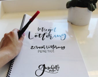 May include: A white notebook with the text "Let's get lettering" and "Brush lettering practice" written in black ink. The notebook also has the text "gmichelle designs" written in black ink. A hand is holding a red and black marker over the notebook.