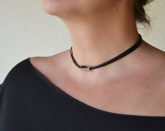 May include: A black choker necklace with a rectangular silver-colored pendant. The necklace is worn around the neck. The person is wearing a black top.
