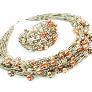 May include: A necklace and bracelet set. The jewellery features multiple strands of silver-coloured cord, decorated with peach, cream, and gold-coloured faux pearls. The set is displayed against a white background.
