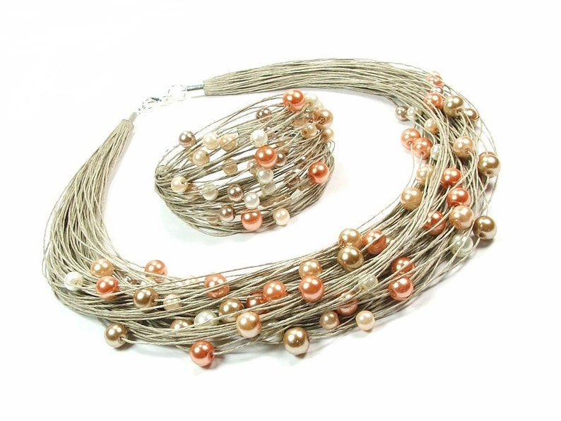 May include: A necklace and bracelet set. The jewellery features multiple strands of silver-coloured cord, decorated with peach, cream, and gold-coloured faux pearls. The set is displayed against a white background.
