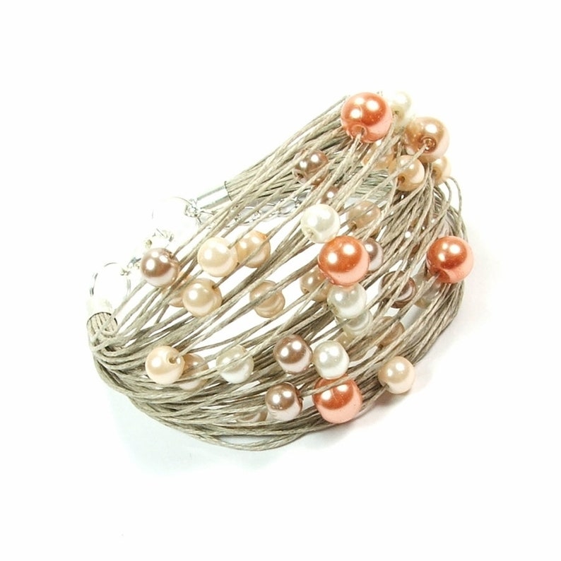 May include: A multi-strand bracelet with a woven, light-coloured base. It is decorated with faux pearls in white, peach, and brown. The bracelet has a silver-coloured clasp.
