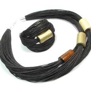 Könnte beinhalten: Ein schwarzes Halsketten- und Armband-Set. Die Halskette besteht aus mehreren Strängen schwarzer Kordel, die mit zylindrischen Holzperlen in Gold und Braun akzentuiert sind. Das Armband hat ein ähnliches Design mit einer einzelnen goldenen Perle. Der Schmuck ist vor weißem Hintergrund.