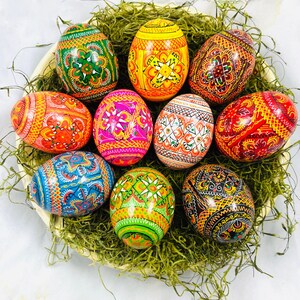 Könnte beinhalten: Eine Sammlung von zehn aufwendig bemalten Ostereiern in verschiedenen Farben und Mustern, darunter Rot, Orange, Gelb, Grün, Blau und Pink. Die Eier sind in einem Nest aus grünem Moos angeordnet.