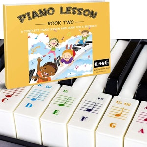 Peut inclure: Un livre jaune intitulé "Piano Lesson Book Two: A Complete Piano Lesson and Guide for a Beginner" avec des enfants dessinés jouant du piano. Le livre est à côté d'un clavier avec des touches blanches et noires et des autocollants colorés avec des notes de musique et des lettres.