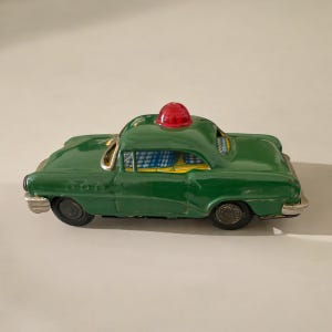 May include: A green toy car with a red light on top. The car has a checkered interior and is made of metal.