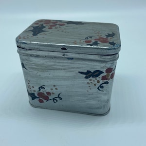 May include: A vintage metal tin with a floral design. The tin is painted in shades of gray, red, and black. The lid has a small hole in the center.