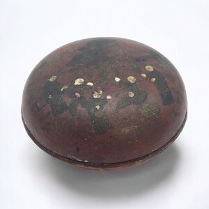May include: A round, reddish-brown container with a lid. The surface has dark, irregular patterns and small, gold-colored accents. The container appears to be made of a leather-like material and has a seam around the middle.