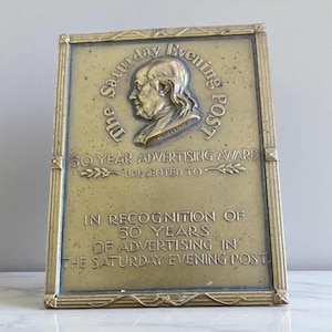May include: A rectangular brass plaque with an antique finish, featuring the text "The Saturday Evening Post" and a profile of a man. The plaque is an advertising award, recognising 50 years of advertising in the publication. The text "50 Year Advertising Award" is visible.