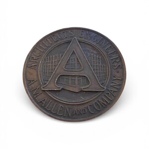 Puede incluir: Un emblema circular de color bronce con el texto "A.M. ALLEN AND COMPANY" y "ARCHITECTS ENGINEERS". El dise&ntilde;o central presenta una "A" estilizada y elementos arquitect&oacute;nicos, todo en bronce.
