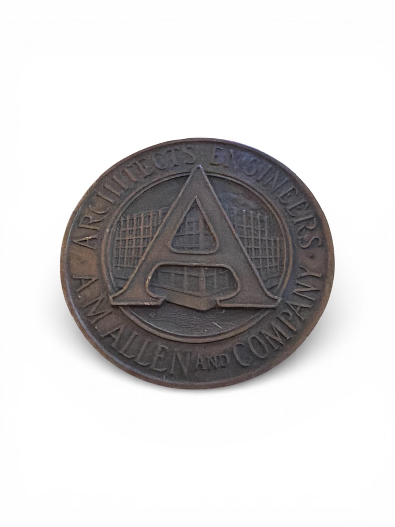 Puede incluir: Un emblema circular de color bronce con el texto "A.M. ALLEN AND COMPANY" y "ARCHITECTS ENGINEERS". El dise&ntilde;o central presenta una "A" estilizada y elementos arquitect&oacute;nicos, todo en bronce.