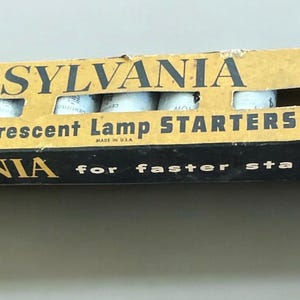 Puede incluir: Una caja vintage de arrancadores de lámparas fluorescentes Sylvania. La caja está amarillenta por la edad y presenta el logotipo y el texto de Sylvania. La caja tiene una sección recortada que revela los arrancadores en el interior. El texto "for faster starting!" está impreso en la caja.