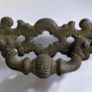 May include: A vintage, ornate, brass door pull with a decorative, openwork design. The pull is in a worn, aged finish.