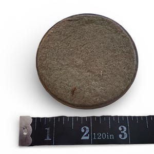 Puede incluir: Un objeto redondo cubierto de fieltro con un borde marr&oacute;n oscuro. Una cinta m&eacute;trica negra se coloca debajo, mostrando medidas de hasta 7,6 cm.