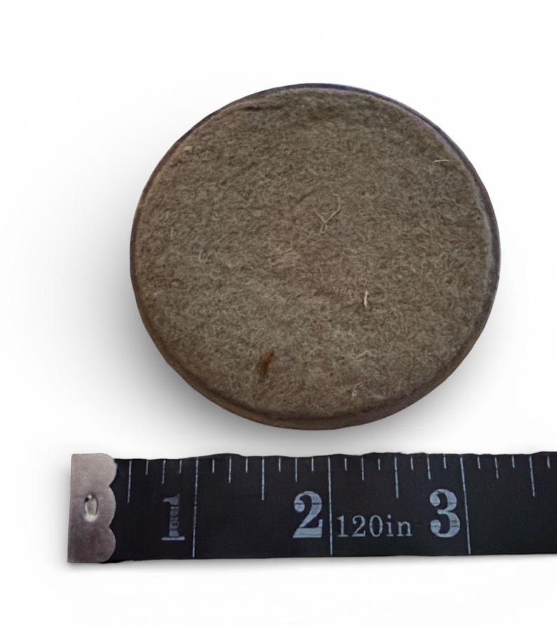 Puede incluir: Un objeto redondo cubierto de fieltro con un borde marr&oacute;n oscuro. Una cinta m&eacute;trica negra se coloca debajo, mostrando medidas de hasta 7,6 cm.