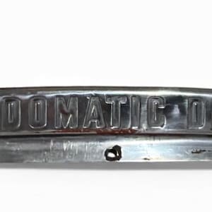 May include: A vintage chrome "FORDOMATIC DRIVE" emblem. The emblem is rectangular with rounded edges and features raised lettering. The metal has a worn, aged appearance with some rust and a small hole.