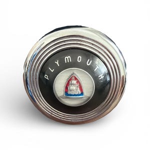 May include: A vintage Plymouth car emblem. The emblem is circular with a chrome finish and a black center. The center features the word "PLYMOUTH" in white lettering and a red, white, and blue ship logo. A classic car part.
