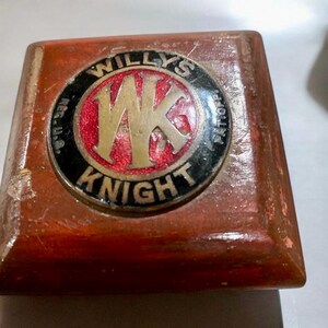Può includere: Una scatola di legno con un emblema circolare in ottone che presenta il testo "WILLYS KNIGHT" e un logo "WK" stilizzato in oro su sfondo rosso.