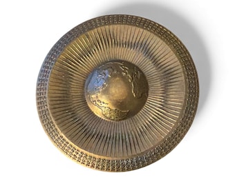 May include: A vintage brass-colored decorative plate featuring a detailed relief of the Earth at the center. The plate has a sunburst design radiating from the center and a patterned border with the numbers 1931-1944. The plate is on a white background.
