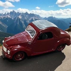 May include: A burgundy-colored vintage toy car with a silver roof, parked on a paved road. The car has a classic design with rounded features and a detailed front grille. The background features a mountain range under a blue sky with clouds.