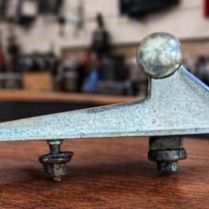 May include: A weathered, silver-coloured metal car component, featuring a spherical top and a triangular base. The part has two threaded studs and is likely an exterior trim piece. It rests on a wooden surface.