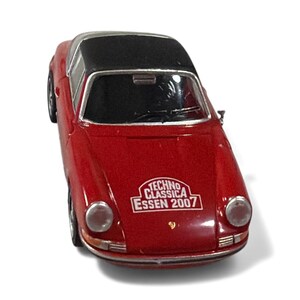 May include: A red toy car with a black roof. The car has a sticker on the front that says "Techno Classica Essen 2007". The car has silver headlights and wheels.