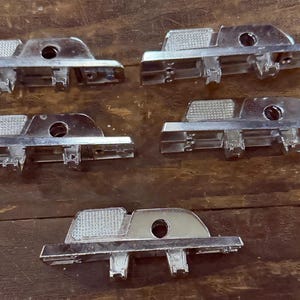 May include: Five chrome-colored automotive trim pieces are arranged on a wooden surface. Each piece has a rectangular base with a curved top, a central hole, and a textured section. The pieces appear to be new.