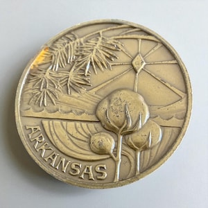 May include: A round, bronze-colored medallion featuring a landscape scene. The word "ARKANSAS" is inscribed at the bottom. The design includes cotton plants, a Ferris wheel, and a tree branch.