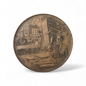 May include: A bronze commemorative coin with detailed relief imagery. The coin features buildings, a horse-drawn carriage, figures, an airplane, a car, and the dates 1854-1954. The coin has a circular shape and a metallic finish.