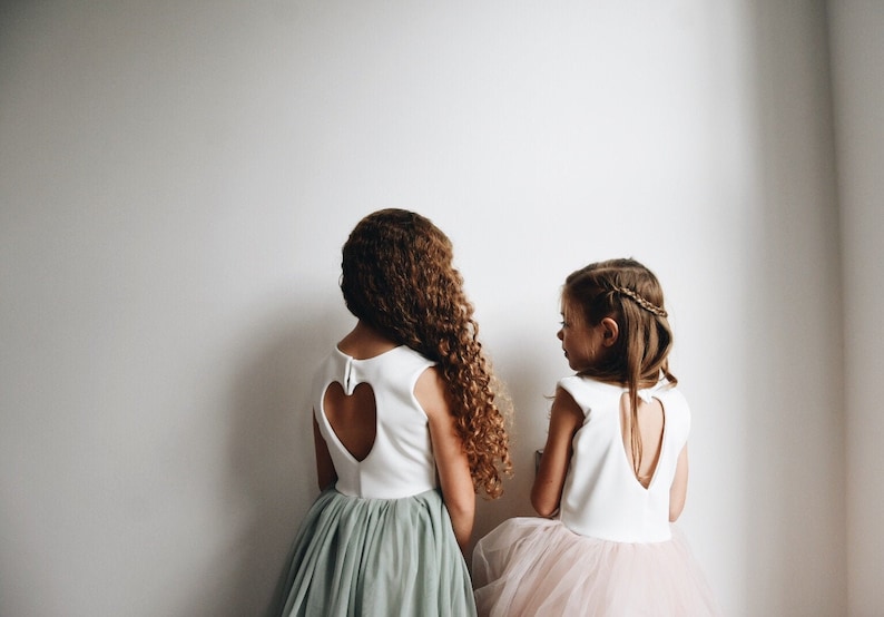May include: Two girls wearing white dresses with heart-shaped cutouts in the back. The girl on the left is wearing a sage green tulle skirt, while the girl on the right is wearing a pink tulle skirt.