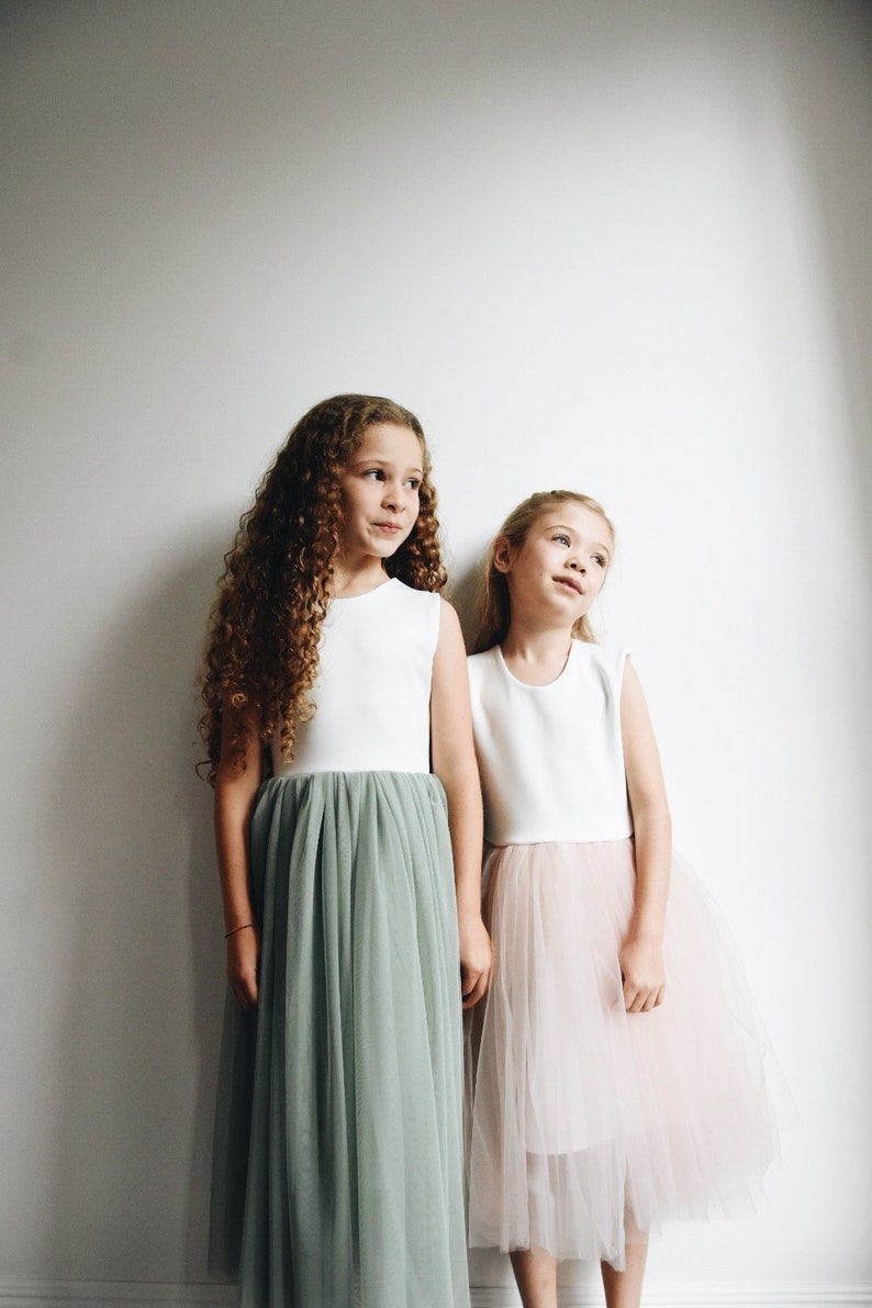 May include: Two girls wearing white sleeveless tops and tulle skirts. The girl on the left is wearing a sage green tulle skirt and has curly brown hair. The girl on the right is wearing a blush pink tulle skirt and has straight blonde hair.