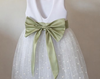 Op de afbeelding: Een mouwloze witte jurk met een lage rug, een saliegroene satijnen strik en een tule rok met kleine, sprankelende accenten. De jurk hangt aan een houten kleerhanger.
