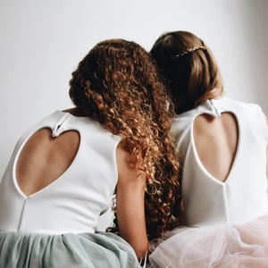 May include: Two girls wearing white dresses with heart-shaped cutouts in the back. The girl on the left has curly brown hair and is wearing a sage green tulle skirt. The girl on the right has straight brown hair and is wearing a pink tulle skirt.