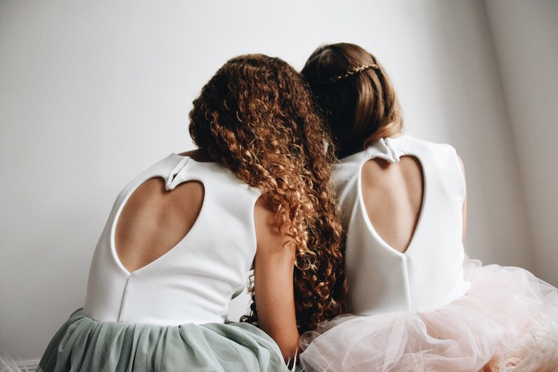 May include: Two girls wearing white dresses with heart-shaped cutouts in the back. The girl on the left has curly brown hair and is wearing a sage green tulle skirt. The girl on the right has straight brown hair and is wearing a pink tulle skirt.