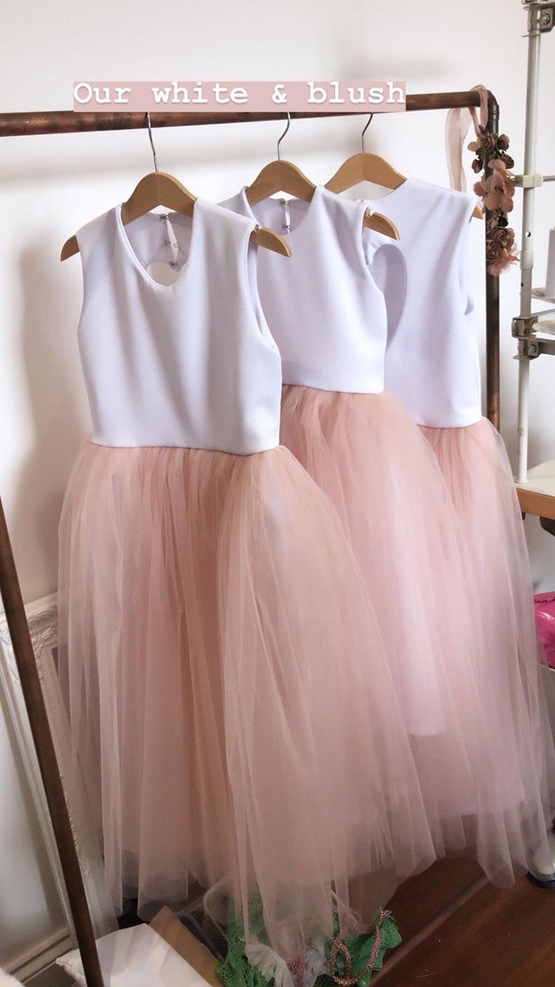 May include: Three white and blush tulle dresses hanging on a rack. The dresses have a white bodice and a blush tulle skirt. The text "Our white & blush" is visible at the top of the image.