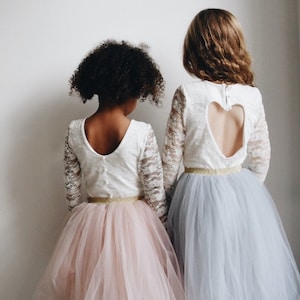 Op de afbeelding: Twee meisjes dragen witte kant tops met lange mouwen en tule rokken. De ene rok is roze en de andere is blauw. De witte top heeft een hartvormige uitsnijding in de rug.